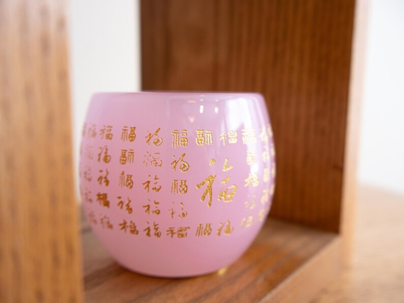 May include: A pink glass votive candle holder with gold Chinese characters. The round candle holder sits on a wooden shelf. The candle holder is a soft pink color.