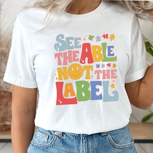 May include: White t-shirt with a colorful retro-style graphic that says "See the able not the label" with a smiley face.