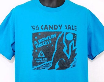 Puede incluir: Una camiseta azul turquesa con un estampado negro de un lobo aullando a la luna. El estampado también incluye el texto "'96 CANDY SALE", "A HOWLIN' SUCCESS" y "Camp Fire Boys and Girls".