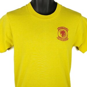 Puede incluir: Camiseta amarilla de manga corta con un parche bordado en rojo en la parte delantera. El parche presenta un tocado de nativo americano y el texto "Hondo Valley" y "Sound Marines".