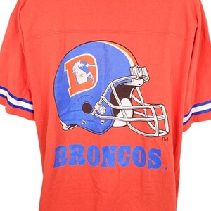 May include: Orange t-shirt featuring a blue and orange football helmet graphic with the word "BRONCOS" in blue. The shirt has white and blue stripes on the sleeves.