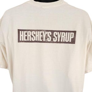 May include: Cream-colored t-shirt featuring the text "HERSHEY'S SYRUP" in a brown rectangular box. The shirt has short sleeves and a crew neck. The text is centered on the chest.