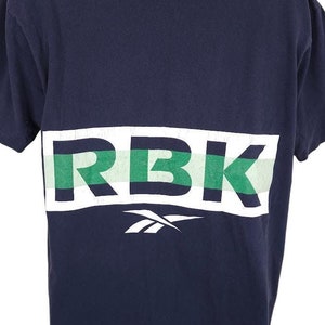 Op de afbeelding: Een marineblauw t-shirt met een wit en groen gestreepte grafische afbeelding met de tekst "RBK" op de rug.