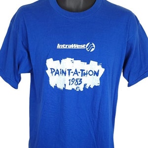 May include: A royal blue t-shirt featuring the text "PAINT-A-THON 1983" in white, and the "IntraWest" logo. The short-sleeved shirt is likely vintage.
