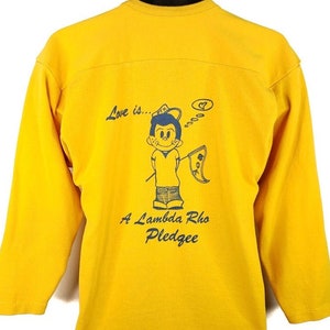 Peut inclure: T-shirt à manches longues jaune avec un personnage de dessin animé portant un chapeau et tenant un drapeau. Le texte "Love is..." et "A Lambda Rho Pledgee" est imprimé sur le t-shirt.