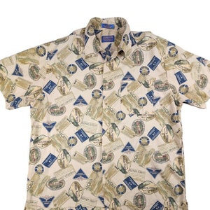 Puede incluir: Una camisa de botones de manga corta beige con un patrón repetido de etiquetas marrones y verdes con la palabra "ALOHA" impresa en ellas. Las etiquetas están impresas sobre un fondo crema.
