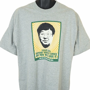 May include: A grey t-shirt with a graphic of a man wearing glasses and the text "STEVE SAYS: DEFEND YOUR OS CHOICE OR YOU WILL LOSE IT. JOIN THE FIGHT FOR THE MAC." The graphic is in green and yellow.