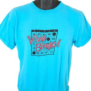 May include: A turquoise blue t-shirt with a black and white bingo card graphic. The card has the word "Viva Bingo!" printed in red and black letters. There are black dots on the card.