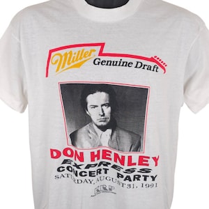 Puede incluir: Una camiseta blanca de manga corta con un estampado gráfico que incluye el logotipo de Miller Genuine Draft en rojo y amarillo. Debajo hay un retrato en blanco y negro de Don Henley, con el texto Don Henley Express Concert Party Saturday, August 31, 1991.
