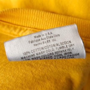 May include: A white care label with black text on a yellow t-shirt. The label reads "Made in U.S.A. Fabrique Aux Etats-Unis Hecho En EE. UU. 100% COTTON COTON/ALGODON MACHINE WASH COLD, TUMBLE DRY LOW, WASH COLORS SEPARATELY. BLEACH. COOL IRON. ALEAU FROIDE PASS"