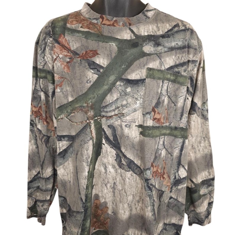Op de afbeelding: Camouflage shirt met lange mouwen, een opstaande kraag en een borstzak. Het shirt heeft een patroon van groen, bruin en grijs, dat lijkt op een bosrijke omgeving. Het shirt wordt gepresenteerd op een zwarte mannequin.