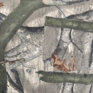 Op de afbeelding: Close-up van een camouflagepatroonstof. Het ontwerp toont een mix van groen, bruin en grijs, die een bosrijke omgeving nabootsen. De stof heeft een zak met een groene rand. De tekst "Mossy Oak Treestand" is zichtbaar.