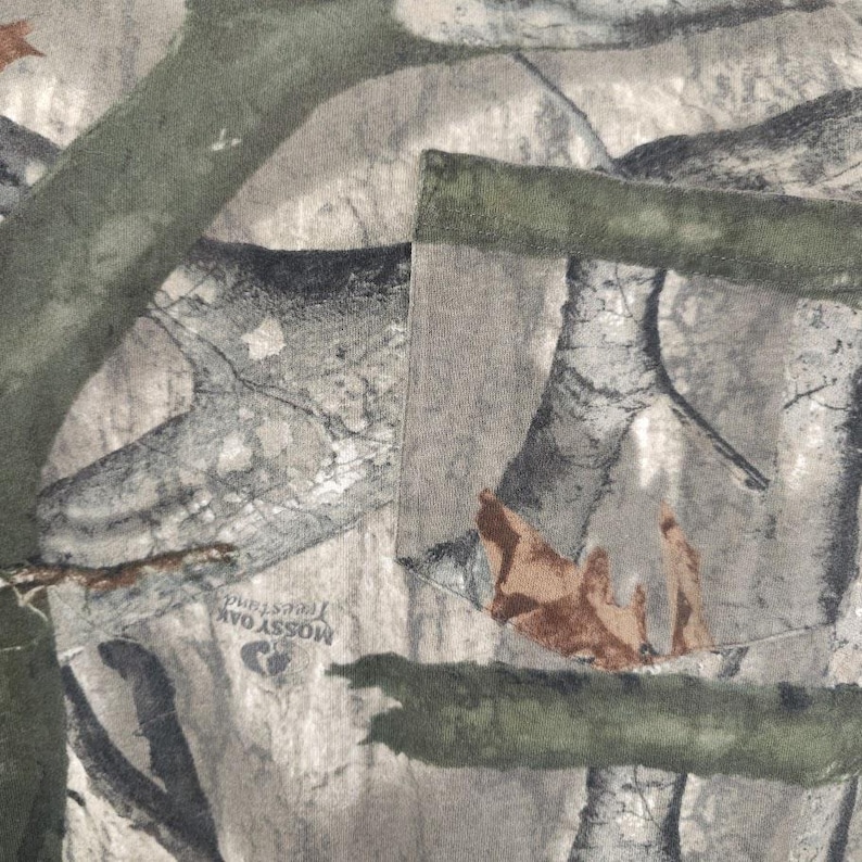 Op de afbeelding: Close-up van een camouflagepatroonstof. Het ontwerp toont een mix van groen, bruin en grijs, die een bosrijke omgeving nabootsen. De stof heeft een zak met een groene rand. De tekst "Mossy Oak Treestand" is zichtbaar.