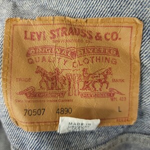 May include: A brown leather Levi Strauss & Co. patch with red stitching. The patch has the company's logo, "Original riveted quality clothing", "Trade Mark", and "Care Instructions Inside Garment". The patch also has the numbers "70507" and "4890" and the letter "L".