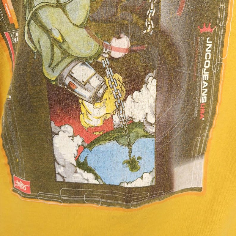 May include: A yellow t-shirt featuring a graphic print of a robot holding a chain connected to a globe. The graphic is within a black frame with the text "JNCO JEANS USA" in white.