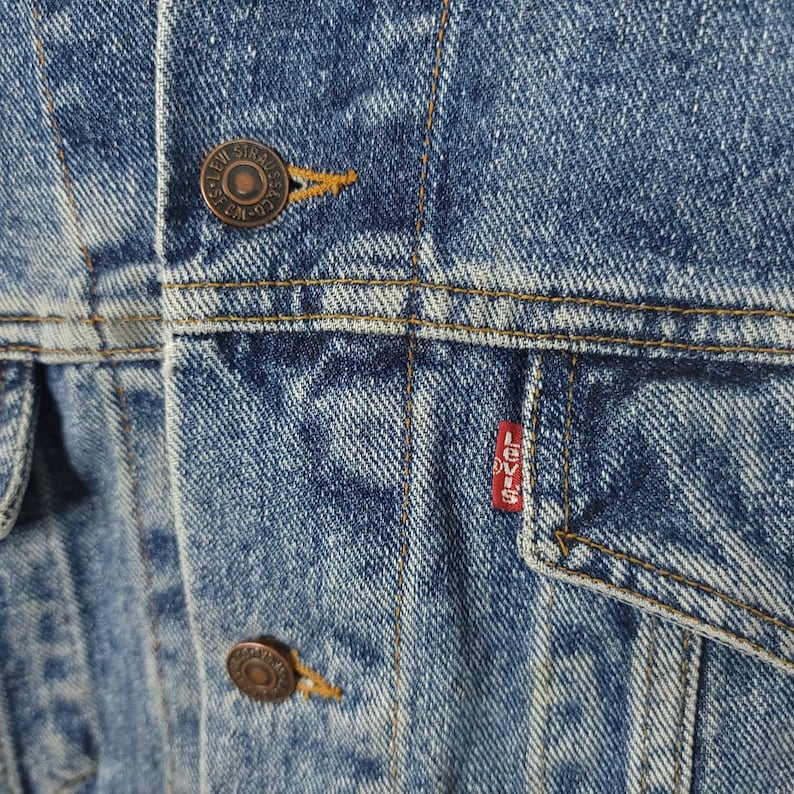 May include: Close-up of a blue denim jacket with a copper button and a red Levi's patch. The denim has a faded, worn look.