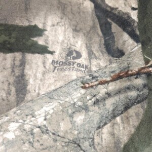 Op de afbeelding: Close-up van camouflage stof met het "Mossy Oak Treestand" logo. De stof heeft een mix van groen, bruin en grijs, die een bosrijke omgeving imiteert. Het logo is donkergrijs.