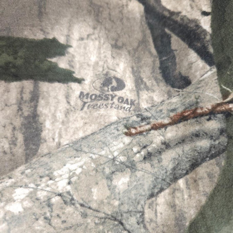 Op de afbeelding: Close-up van camouflage stof met het "Mossy Oak Treestand" logo. De stof heeft een mix van groen, bruin en grijs, die een bosrijke omgeving imiteert. Het logo is donkergrijs.