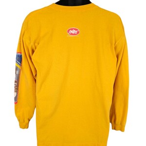 May include: A yellow long-sleeved t-shirt with a red oval logo on the back that says "DVS Shoes". The left sleeve has a colourful graphic design.