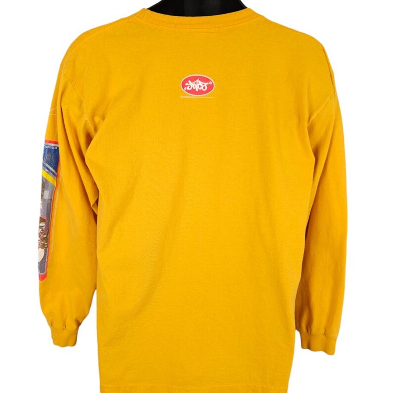 May include: A yellow long-sleeved t-shirt with a red oval logo on the back that says "DVS Shoes". The left sleeve has a colourful graphic design.