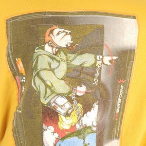 May include: A yellow t-shirt featuring a graphic print of a cartoon character wearing a green hoodie and holding a chain. The character is standing on a globe with a red, yellow and blue design. The text "INCQUEANS" is printed on the shirt.