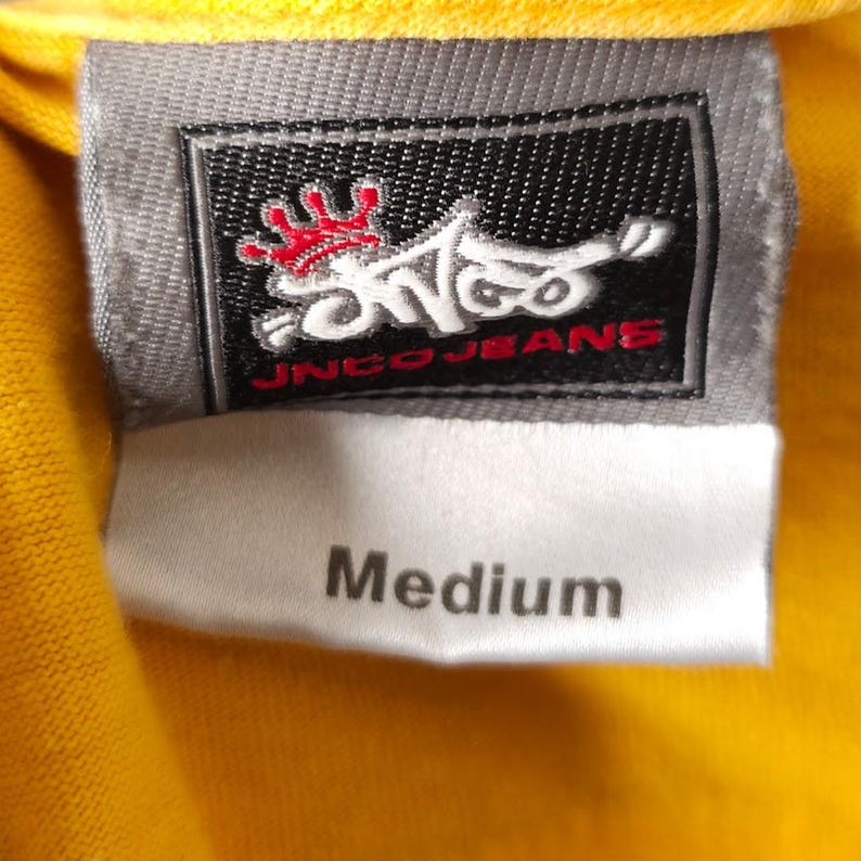 May include: A white tag with black text that reads "Medium" is attached to a yellow garment. The tag is attached to a grey tag with a black and red logo that reads "Inco Jeans".