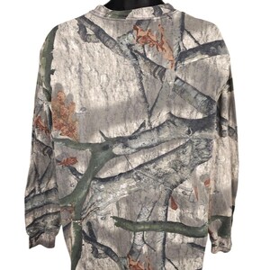 Op de afbeelding: Camouflage shirt met lange mouwen in tinten grijs, bruin en groen. Het shirt heeft een ronde hals en een patroon dat lijkt op boomtakken en bladeren. De stof lijkt een lichtgewicht materiaal te zijn.