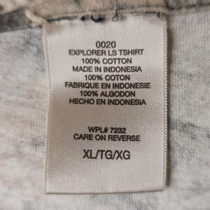Op de afbeelding: Een wit label met zwarte tekst, met "0020 EXPLORER LS TSHIRT 100% COTTON MADE IN INDONESIA." Het label toont ook de maat "XL/TG/XG" en "CARE ON REVERSE."