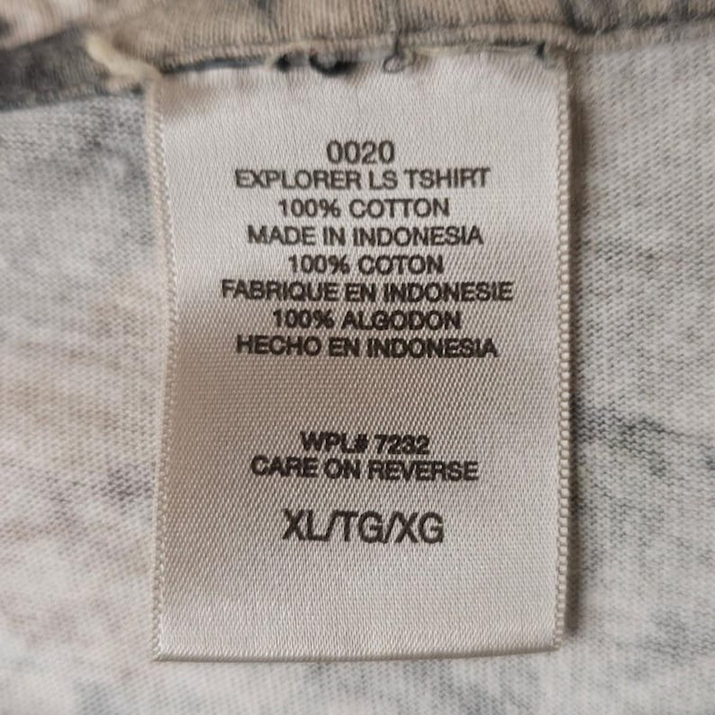 Op de afbeelding: Een wit label met zwarte tekst, met "0020 EXPLORER LS TSHIRT 100% COTTON MADE IN INDONESIA." Het label toont ook de maat "XL/TG/XG" en "CARE ON REVERSE."