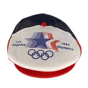 May include: A white and blue baseball cap with a red star and the text "Los Angeles 1984 Olympics" and five Olympic rings.