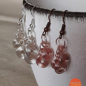 Può includere: Primo piano di quattro paia di orecchini fatti a mano con filo metallico. Due paia sono argentati e due color rame. Ogni orecchino presenta un design a spirale. Gli orecchini sono appesi davanti a una tazza di ceramica bianca.