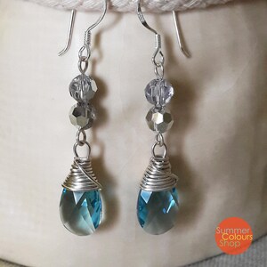 Op de afbeelding: Zilverkleurige oorhangers met een druppelvormige, gefacetteerde blauwe edelsteen. De oorbellen hebben heldere en zilveren kralen en zijn opgehangen aan een zilveren haak.