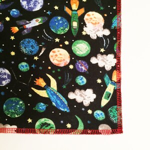 May include: Black fabric with a repeating pattern of colorful rockets, planets, and stars. The fabric has a red stitched edge.