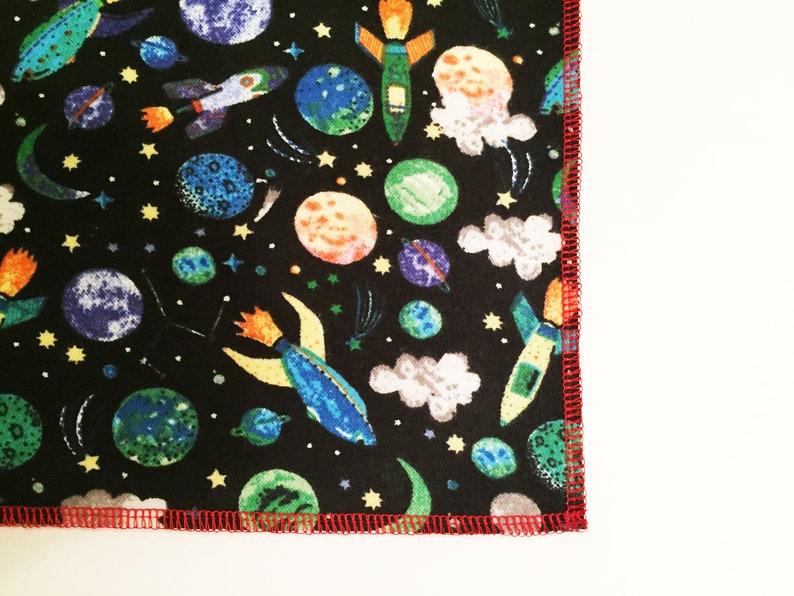 May include: Black fabric with a repeating pattern of colorful rockets, planets, and stars. The fabric has a red stitched edge.