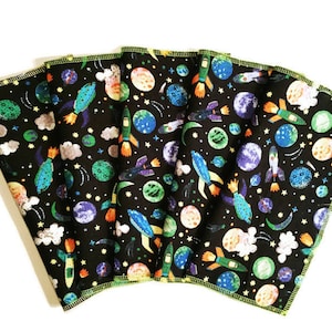 May include: Six black fabric cloths with a colorful space-themed print. The print features planets, rockets, stars, and clouds. The cloths have a yellow border.