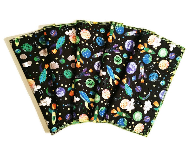 May include: Six black fabric cloths with a colorful space-themed print. The print features planets, rockets, stars, and clouds. The cloths have a yellow border.