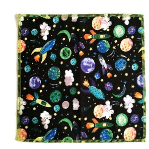 May include: A black fabric square with a colorful space-themed print. The print features rockets, planets, stars, and clouds. The fabric has a green border.