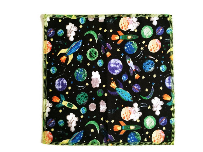 May include: A black fabric square with a colorful space-themed print. The print features rockets, planets, stars, and clouds. The fabric has a green border.