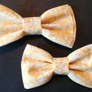May include: Two yellow bow ties with a white floral pattern. The bow ties are made of fabric and have a classic design.