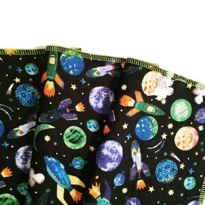 May include: A black fabric with a colorful space-themed print. The print features rockets, planets, and stars. The fabric has a green border.