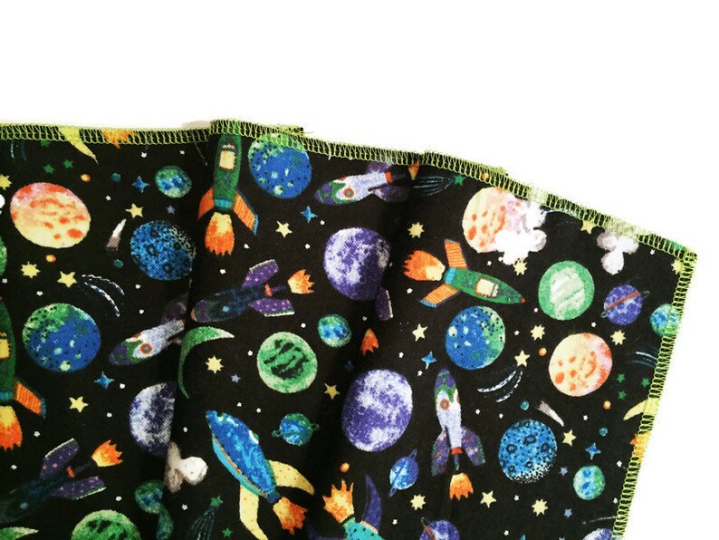 May include: A black fabric with a colorful space-themed print. The print features rockets, planets, and stars. The fabric has a green border.