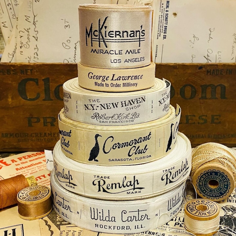 Puede incluir: Una pila de carretes de cinta de tela blanca vintage con texto negro. Los carretes est&aacute;n etiquetados con los nombres de diferentes empresas y ubicaciones, que incluyen "McKiernan's Miracle Mile Los Angeles", "George Lawrence Made to Order Millinery", "The N.Y.-New Haven Shop Robert Kirk, Ltd. San Francisco", "Cormorant Club Sarasota, Florida", "Trade Remlah Mark" y "Wilda Carter Rockford, Ill."