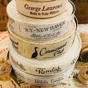 Puede incluir: Una pila de cinco carretes de cinta de tela blanca con etiquetas impresas en negro. Las etiquetas dicen "George Lawrence Made to Order Millinery", "The N.Y.-New Haven Shop Robert Kirk, Ltd. San Francisco", "Cormorant Club Sarasota, Florida", "Trade Mark Remlap" y "Wilda Carter Rockford, Ill."