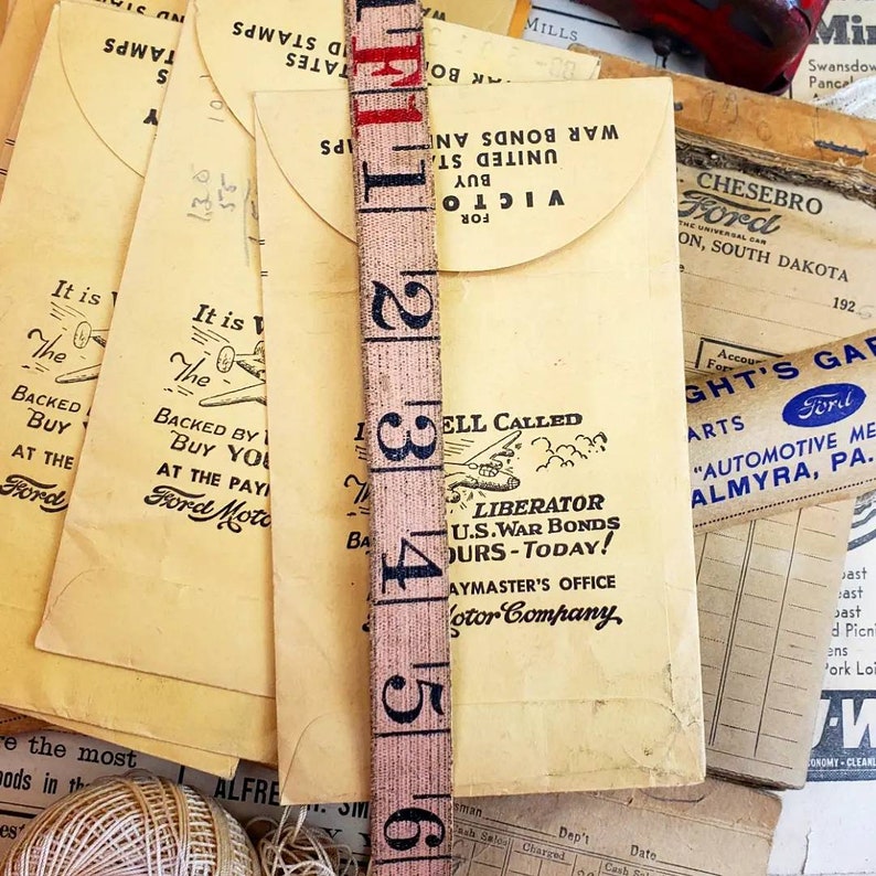 Może przedstawiać: Kolekcja vintage'owych kopert z niebiesko-białym obrazkiem samolotu i tekstem "It is the...Backed by Buy You at the Pay...Ford Motor" oraz "Liberator U.S. War Bonds Ours-Today! Paymaster's Office Motor Company". Koperty są ułożone na vintage'owej taśmie mierniczej z czerwonymi i czarnymi numerami.