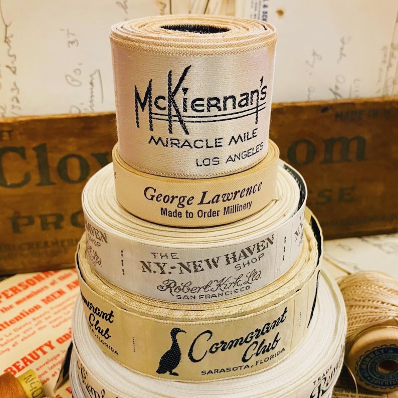 Puede incluir: Una pila de carretes de cinta vintage con etiquetas. El carrete superior tiene una etiqueta que dice "McKiernan's Miracle Mile Los Angeles". El siguiente carrete hacia abajo tiene una etiqueta que dice "George Lawrence Made to Order Millinery". El tercer carrete hacia abajo tiene una etiqueta que dice "The NY-New Haven Shop Robert Kirk, Ltd. San Francisco". El carrete inferior tiene una etiqueta que dice "Cormorant Club Sarasota, Florida".