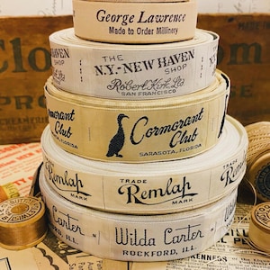 Puede incluir: Una pila de carretes de cinta de tela vintage con diferentes etiquetas. El carrete superior tiene el texto "George Lawrence Made to Order Millinery". El segundo carrete tiene el texto "The N.Y.-New Haven Shop Robert Kirk, Ltd. San Francisco". El tercer carrete tiene el texto "Cormorant Club Sarasota, Florida". El cuarto carrete tiene el texto "Trade Remlah Mark". El quinto carrete tiene el texto "Wilda Carter Rockford, Ill."