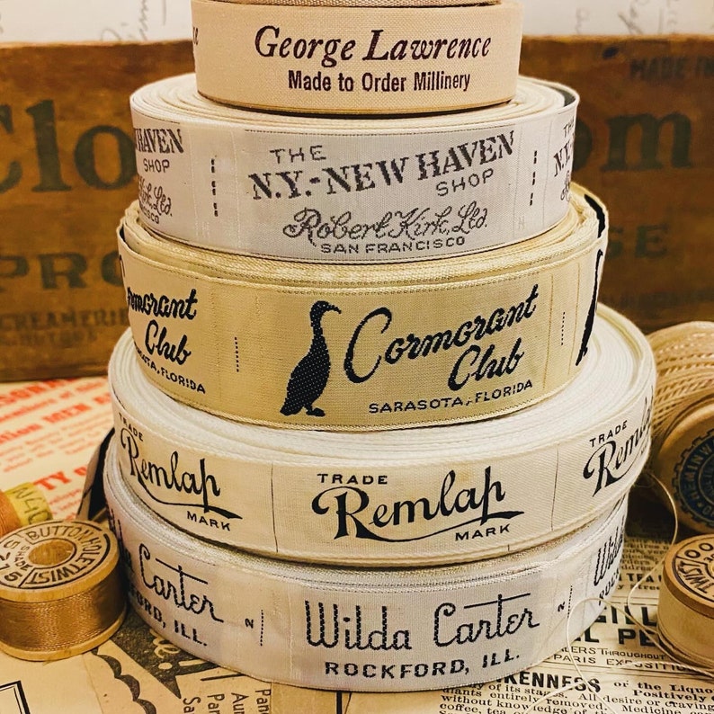 Puede incluir: Una pila de carretes de cinta de tela vintage con diferentes etiquetas. El carrete superior tiene el texto "George Lawrence Made to Order Millinery". El segundo carrete tiene el texto "The N.Y.-New Haven Shop Robert Kirk, Ltd. San Francisco". El tercer carrete tiene el texto "Cormorant Club Sarasota, Florida". El cuarto carrete tiene el texto "Trade Remlah Mark". El quinto carrete tiene el texto "Wilda Carter Rockford, Ill."