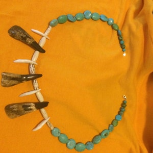 Puede incluir: Un collar de turquesa y hueso con cinco colgantes de dientes de hueso. El collar está sobre una superficie amarilla.