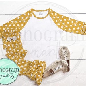 May include: A white and mustard yellow raglan shirt with white polka dots on the sleeves. The shirt has a ruffled trim on the sleeves and bottom. The shirt is paired with mustard yellow pants with white polka dots and a ruffled trim at the bottom. The pants have a white bow on the front. The outfit is laid out on a white wooden surface with two pairs of white shoes.