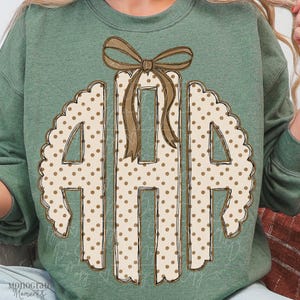 May include: A sage green crewneck sweatshirt featuring a large, centered graphic of the letters A, H, and A in a scalloped circular frame. The letters are cream-colored with brown polka dots and topped with a brown ribbon bow illustration.